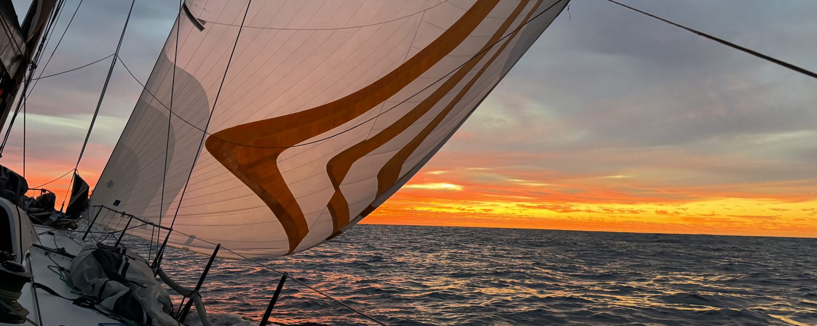 A sailboat under way at sunset, sail carrying orange stripes, open horizon ahead